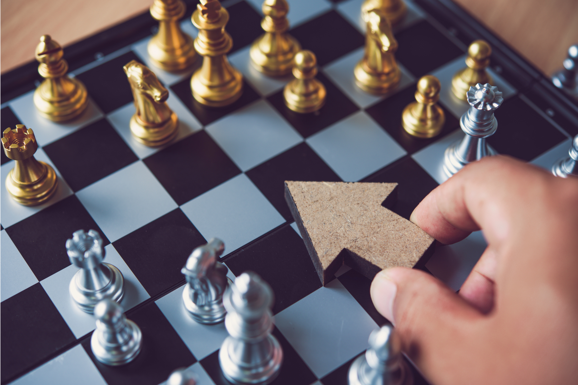 Obrazek dekoracyjny. Plansza do gry w szachy z figurami oraz ręka przesuwająca drewnianą strzałkę symbolizującą kierunek strategii.