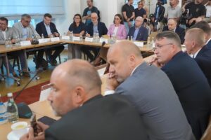 Zdjęcie z rozmów. Ludzie siedzący przy stole. W tle dziennikarze. Wśród siedzących Ałła Witwicka-Dudek oraz Krzysztof Piontek.