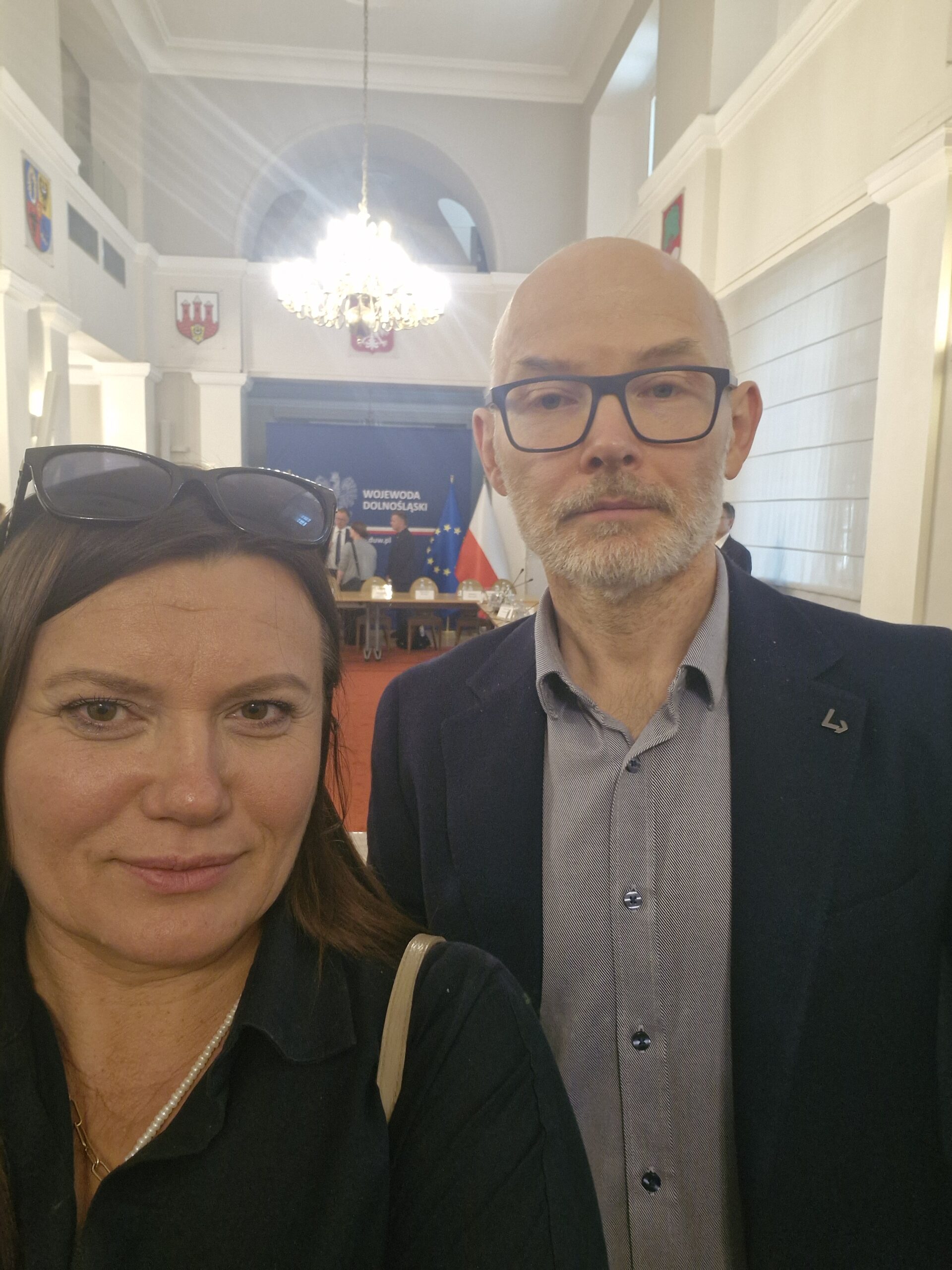 Zdjęcie mgr Ałła Witwicka-Dudek oraz dr hab. inż. Krzysztof Piontek, prof. UEW. Zdjęcie ze spotkania. W tle Sala obrad oraz baner z napisem Wojewoda Dolnośląski na granatowym tle oraz flagi - Polska i Unijna.