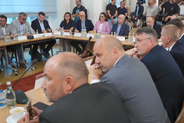 Zdjęcie z rozmów. Ludzie siedzący przy stole. W tle dziennikarze. Wśród siedzących Ałła Witwicka-Dudek oraz Krzysztof Piontek.