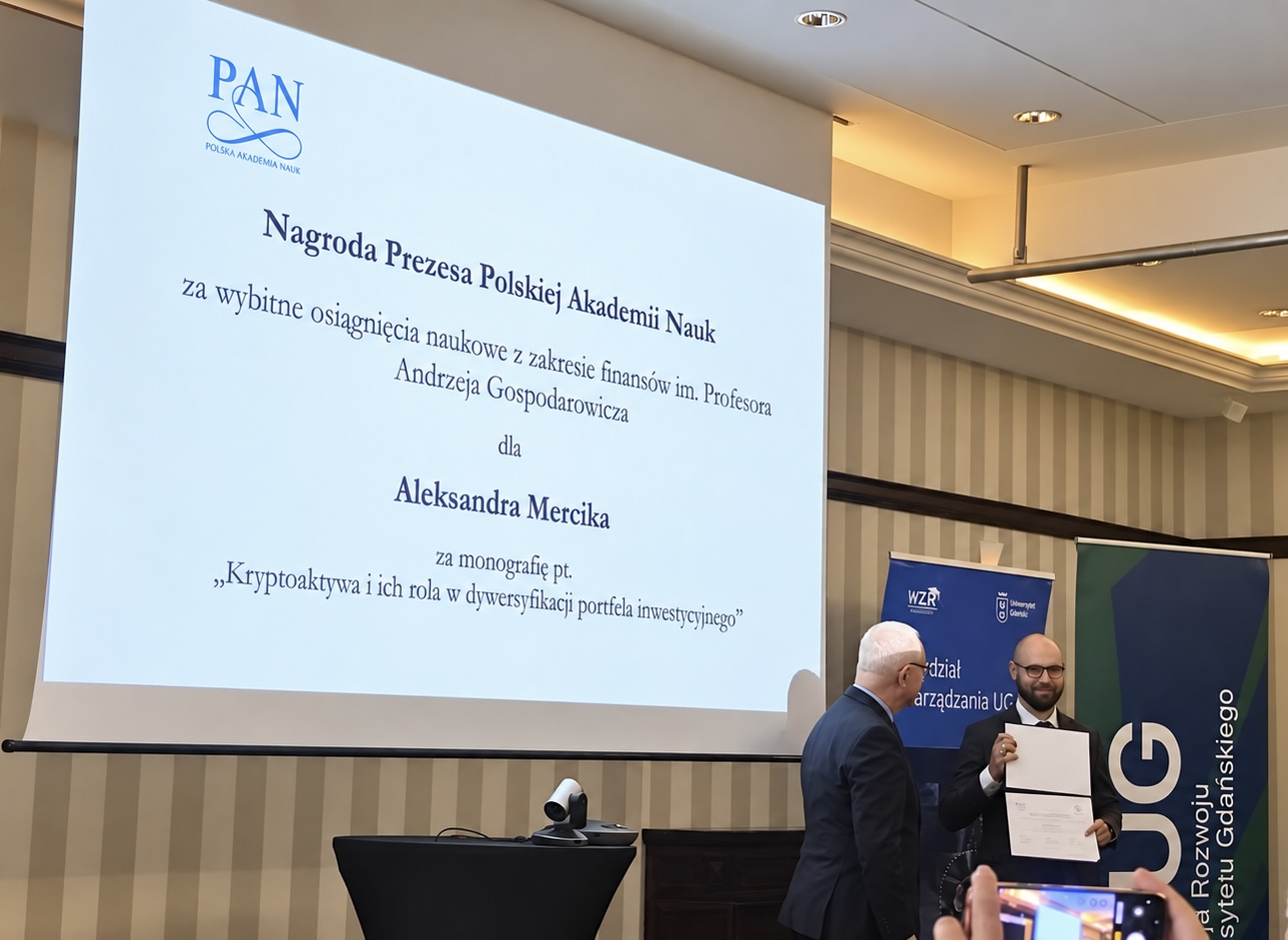 Obraz przedstawia wręczenie nagrody Prezesa PAN. Po lewej stronie wyświetlone na rzutniku napisy: Nagroda Prezesa Polskiej Akademii Nauk za wybitne osiągnięcia naukowe w zakresie finansów im. Profesora Andrzeja Gospodarowicza dla Aleksandra Marcika za monografię pt. "Kryptoaktywa i ich rola w dywersyfikacji portfela inwestycyjnego". Po prawej stronie Aleksander Mercik trzymający dyplom.